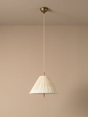 A modern pendant light with a pleated white fabric shade and a brass ceiling canopy, shown hanging against a minimalist beige wall and ceiling.