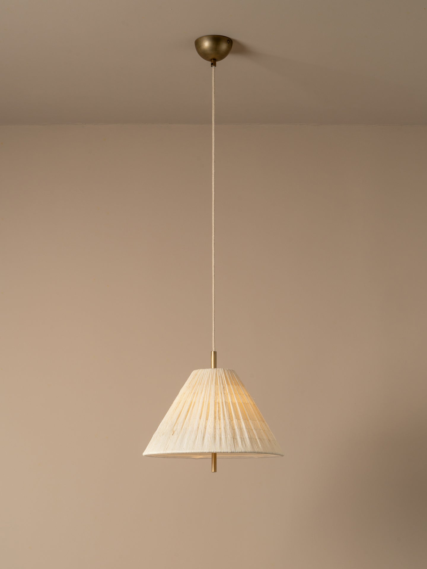 A modern pendant light with a pleated off-white fabric shade and a warm brass metal canopy and cord, shown hanging against a minimalist beige wall and ceiling.