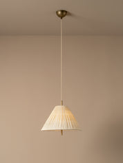 A modern pendant light with a pleated off-white fabric shade and a warm brass metal canopy and cord, shown hanging against a minimalist beige wall and ceiling.