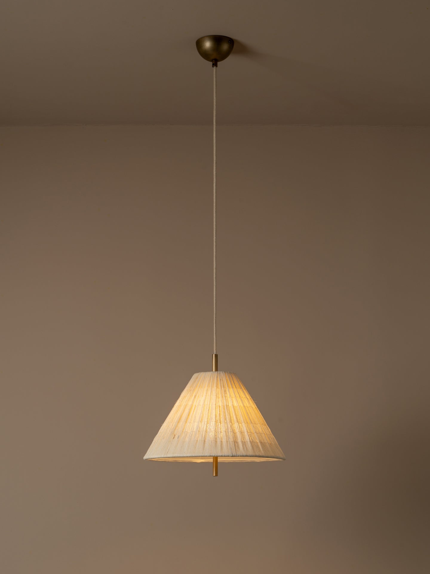 A single pendant light with a pleated beige fabric shade and a dark brass ceiling mount, shown illuminated against a warm taupe wall.