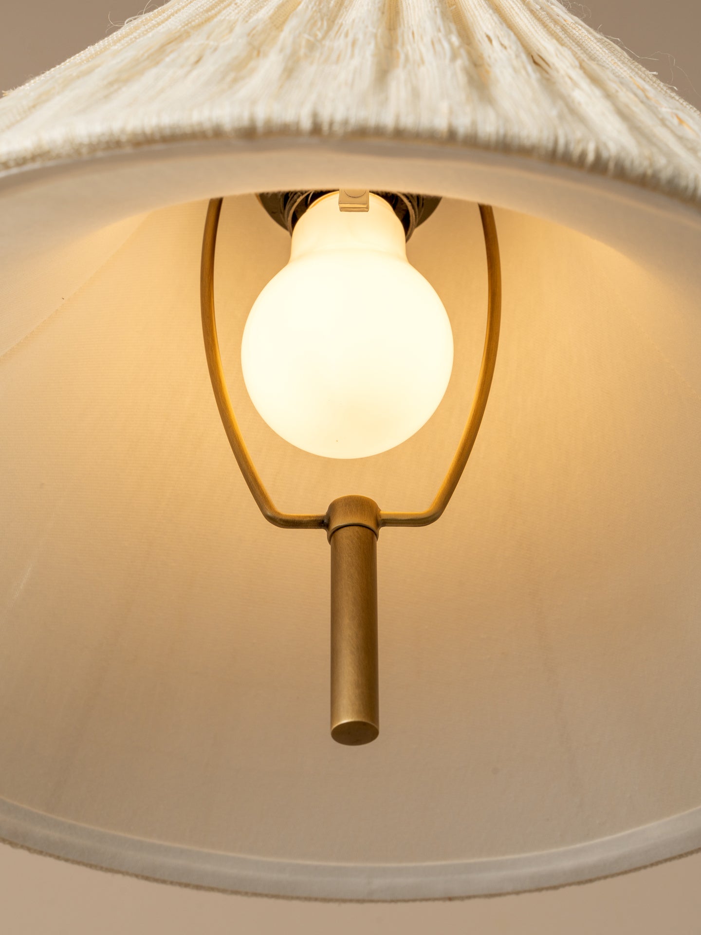 A close-up of a hanging pendant light with a warm white globe bulb glowing inside a brass metal frame, set beneath a textured off-white fabric shade.