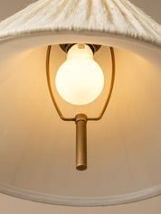 A close-up of a hanging pendant light with a warm white globe bulb glowing inside a brass metal frame, set beneath a textured off-white fabric shade.