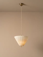 A modern pendant light with a pleated white shade and brass hardware, suspended against a minimalist beige wall and ceiling.