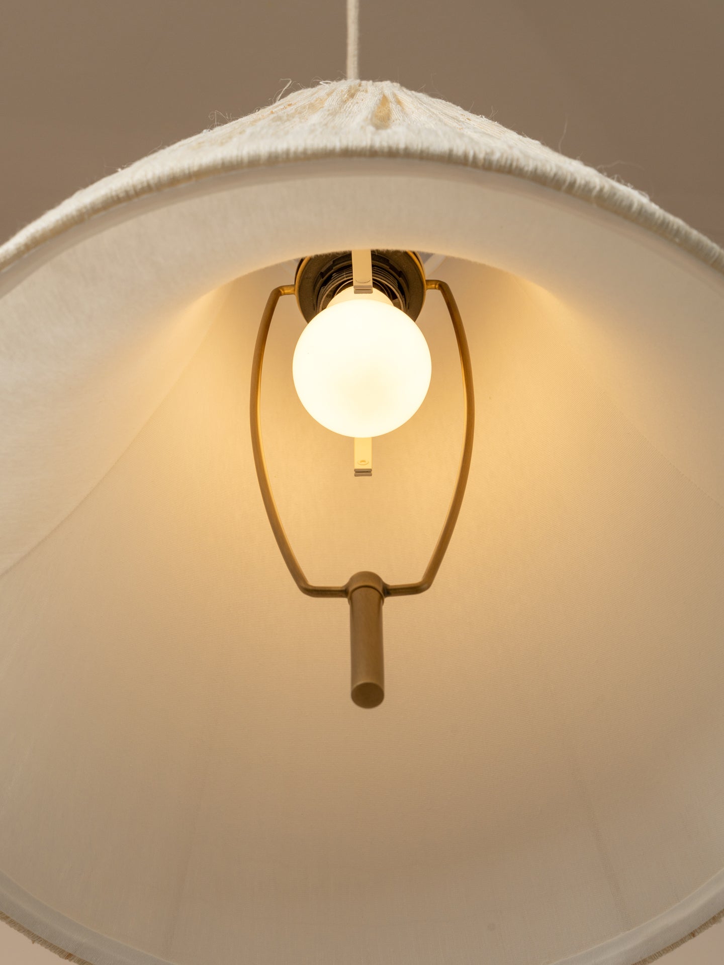 A close-up of a hanging pendant light with a large white fabric drum shade, a glowing warm white globe bulb, and a brass-toned metal frame against a neutral beige background.