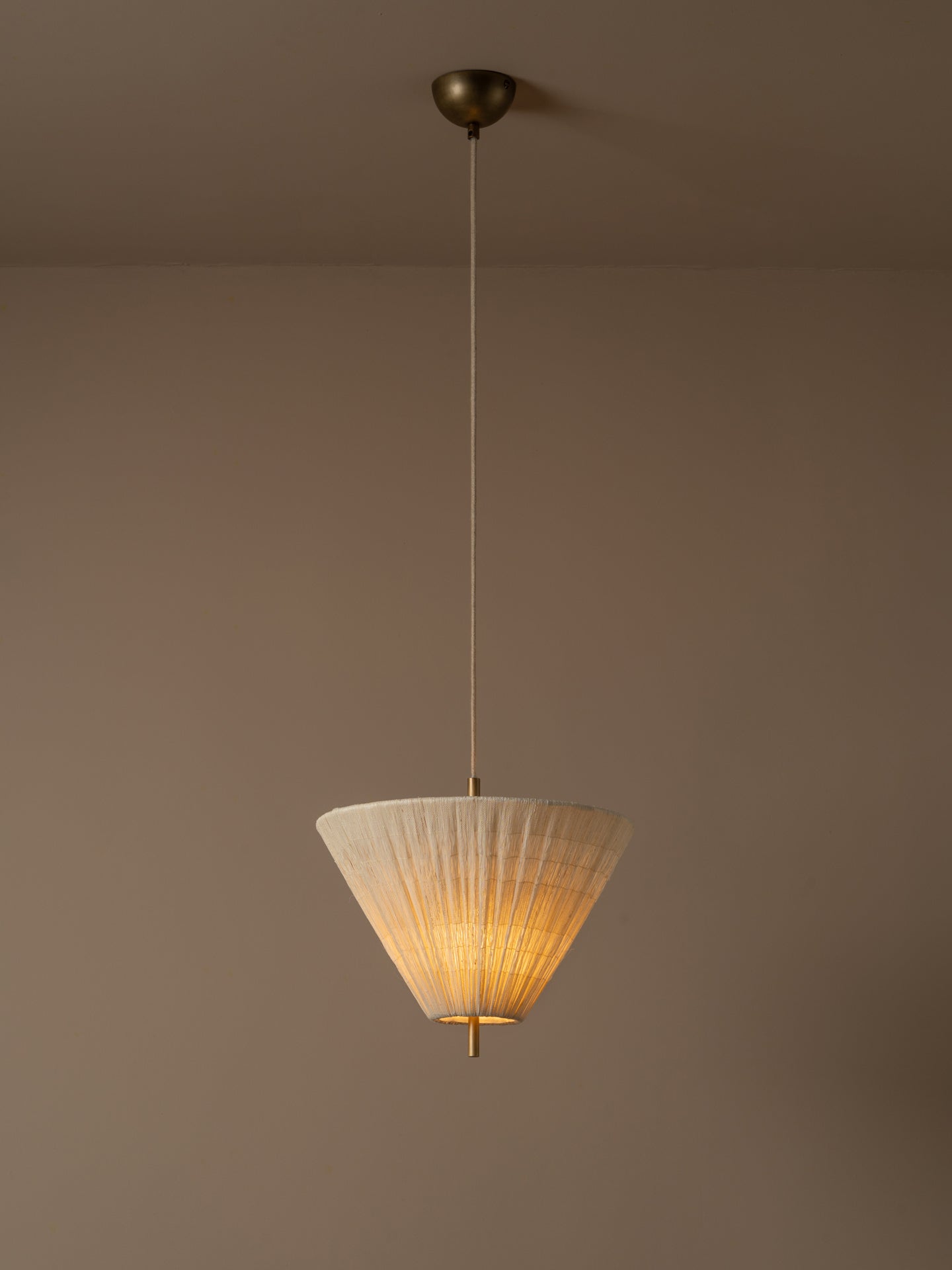 A modern pendant light with a pleated white conical shade and a warm brass ceiling canopy, suspended against a plain taupe wall.