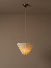 A modern pendant light with a pleated white conical shade and a warm brass ceiling canopy, suspended against a plain taupe wall.