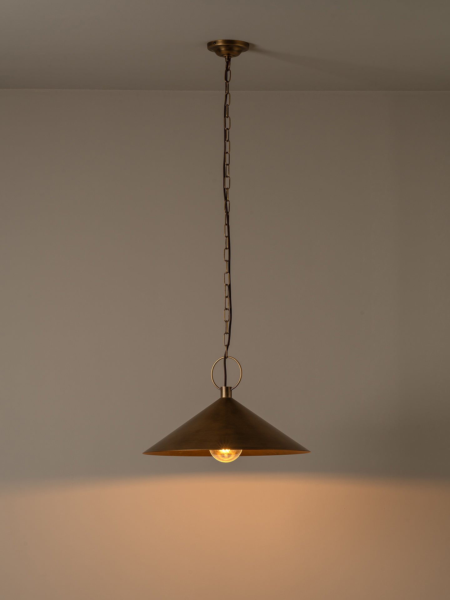 A single brass pendant light with a conical shade hanging from a chain against a plain wall, casting a warm glow downward.