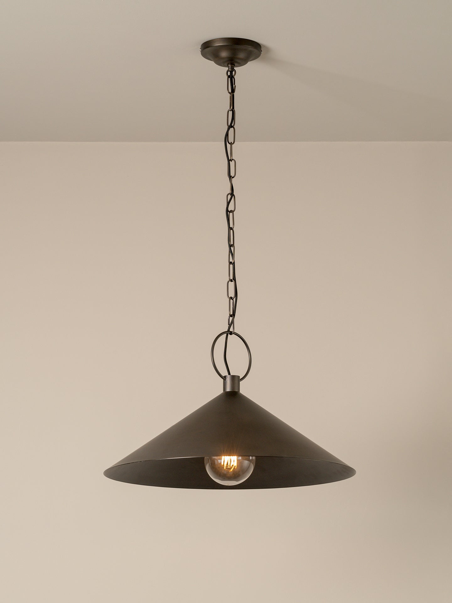 A dark metal pendant light with a wide conical shade hanging from a chain and ceiling canopy against a plain beige wall.