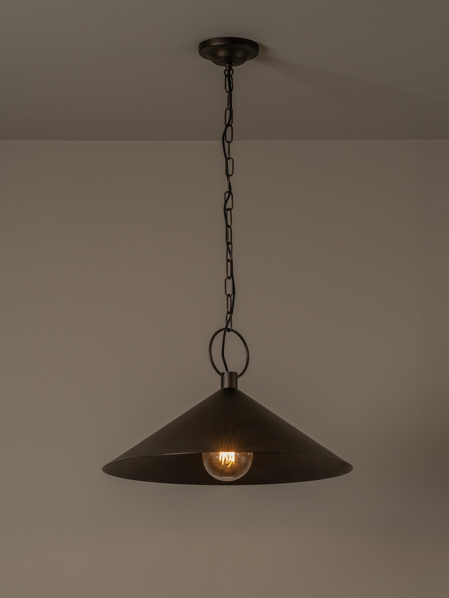A dark metal pendant light with a wide conical shade hanging from a chain and ceiling canopy against a plain beige wall.