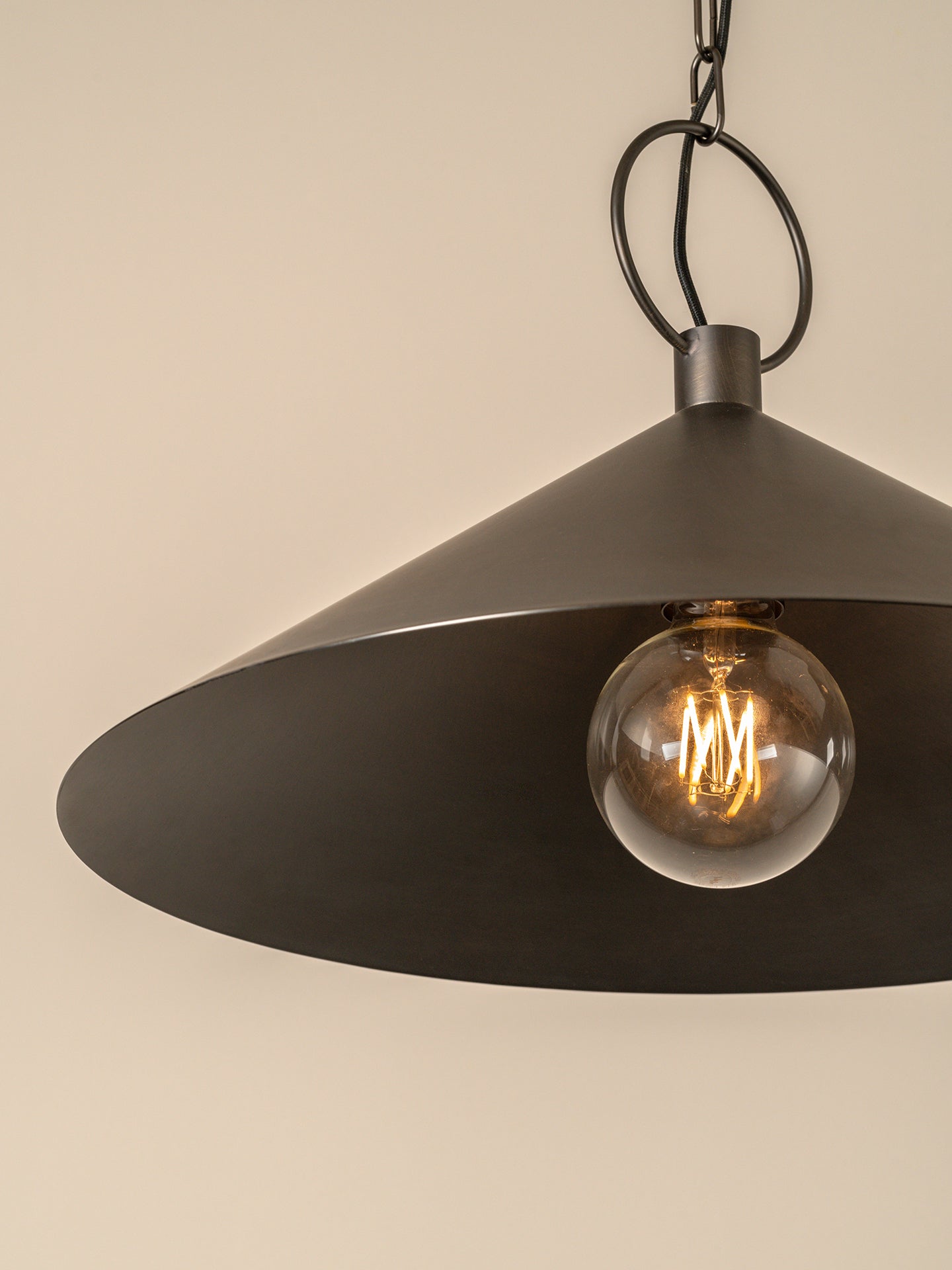 A dark metal pendant light with a wide conical shade hanging from a chain and ceiling canopy against a plain beige wall.