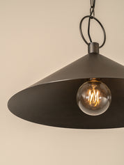 A dark metal pendant light with a wide conical shade hanging from a chain and ceiling canopy against a plain beige wall.