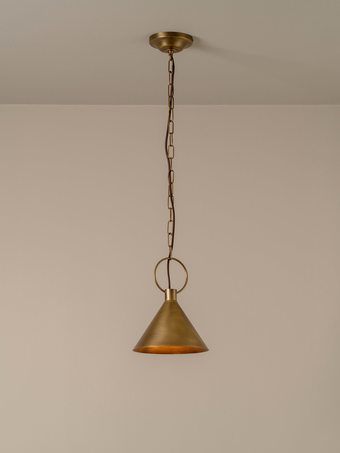 A single brass pendant light hanging from a ceiling with a chain and circular ring, shown against a plain beige wall.