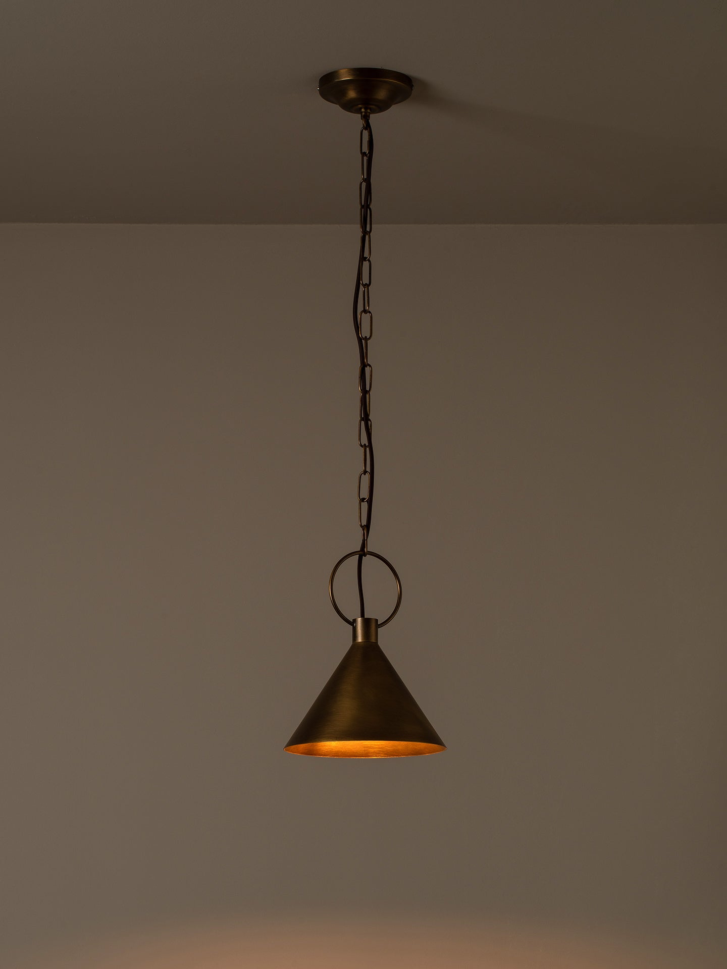A single hanging pendant light with a dark metal chain and ceiling canopy, featuring a conical shade with a warm brass or bronze interior glowing orange, shown against a plain wall.