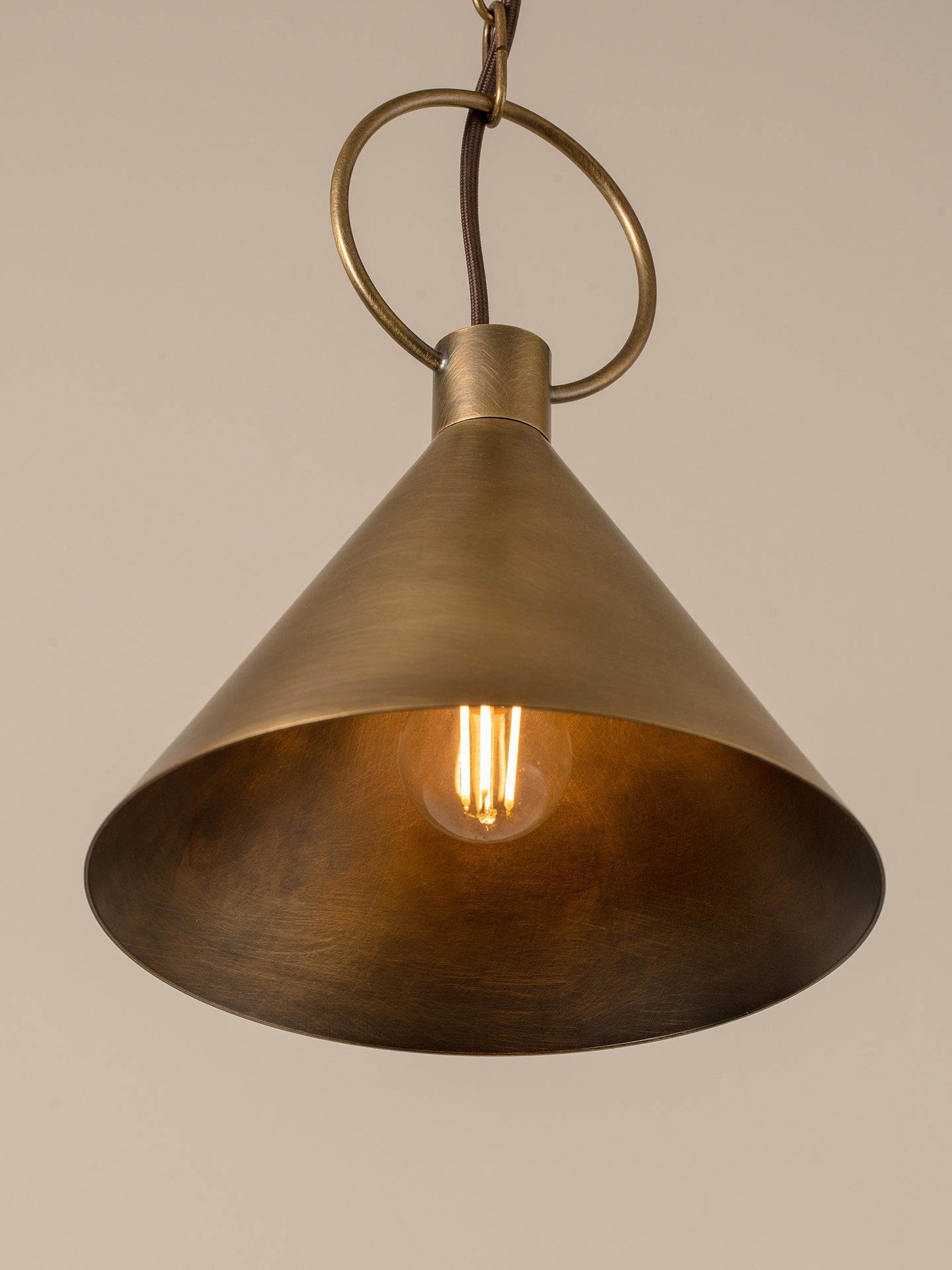 An industrial-style pendant light with a brushed brass cone shade and a visible warm Edison bulb inside, suspended by a circular ring against a plain beige background.