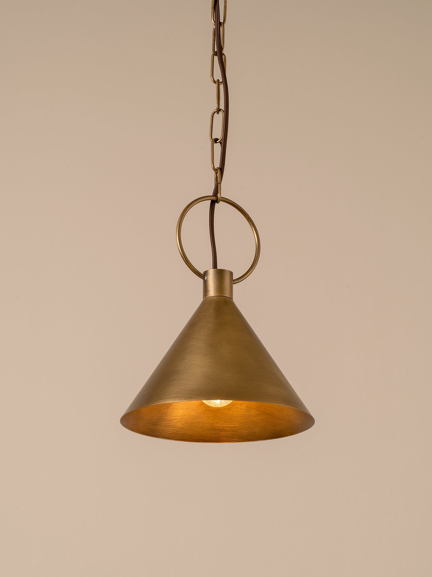 An industrial-style pendant light with a brushed brass cone shade and a visible warm Edison bulb inside, suspended by a circular ring against a plain beige background.