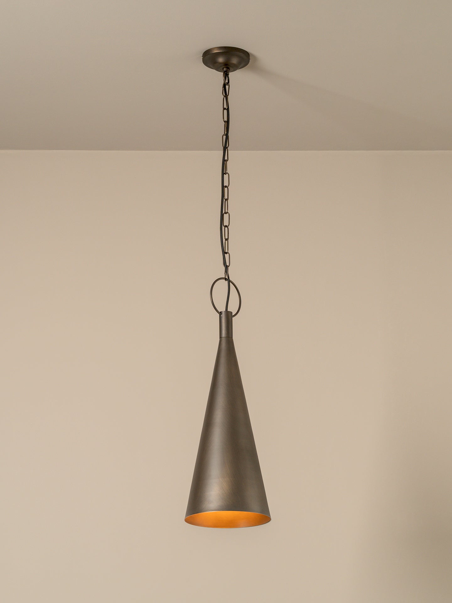 A single pendant light with a dark bronze metal cone shade and a warm orange-gold interior, hanging from a chain against a plain beige wall.