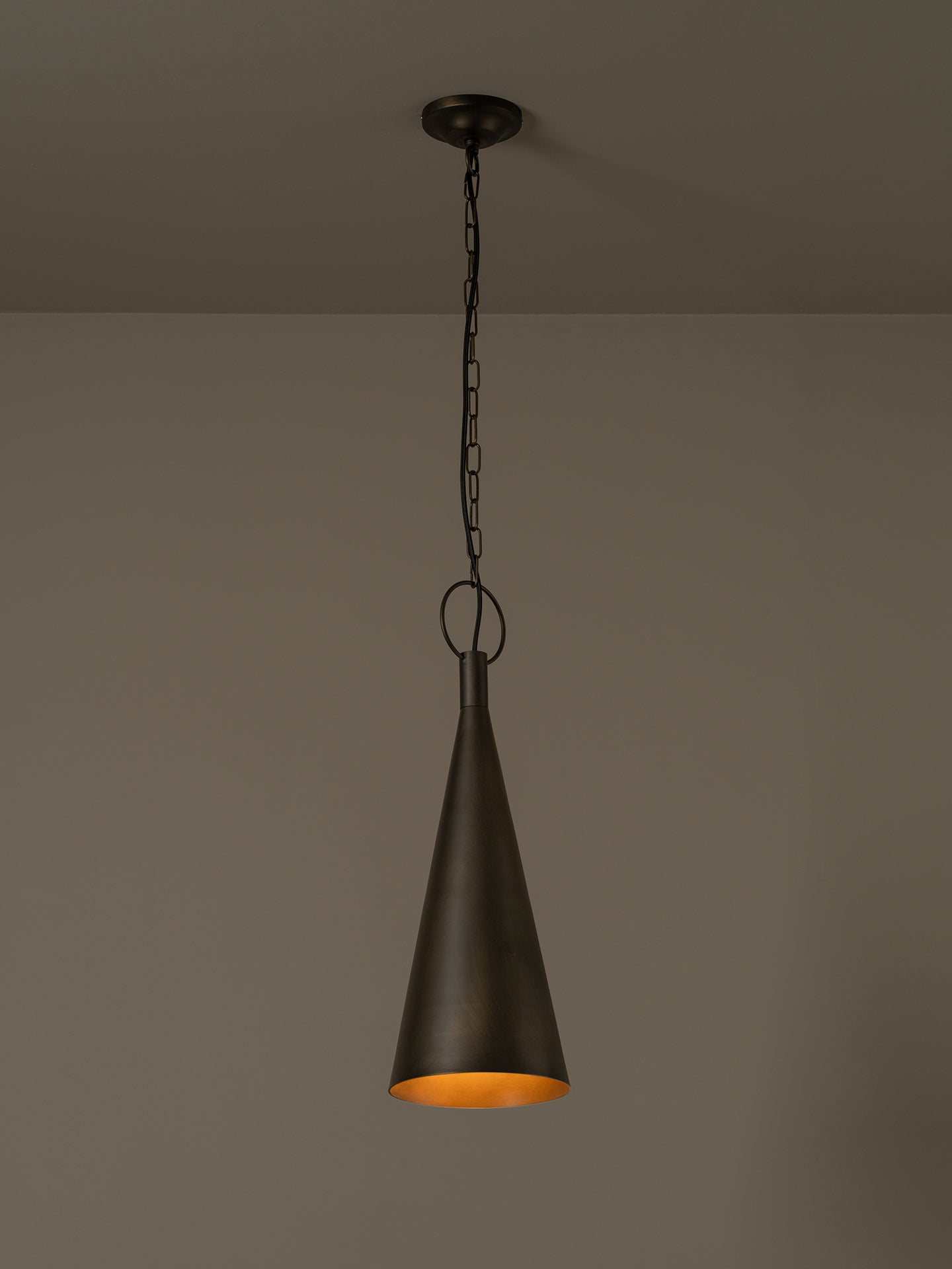 A dark metal pendant light with a tapered cone shade hanging from a chain against a plain taupe wall.