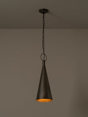 A dark metal pendant light with a tapered cone shade hanging from a chain against a plain taupe wall.
