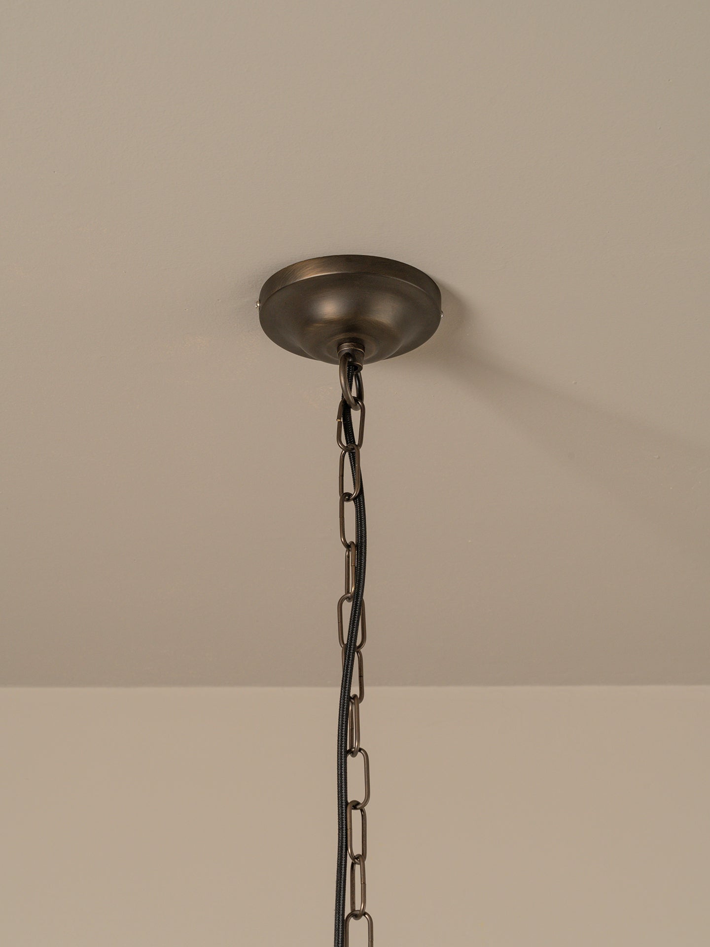 A close-up of a ceiling-mounted pendant light fixture showing a round dark metal ceiling canopy and hanging chain against a plain beige wall and ceiling.