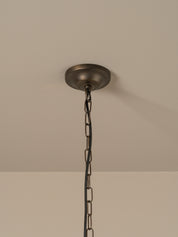 A close-up of a ceiling-mounted pendant light fixture showing a round dark metal ceiling canopy and hanging chain against a plain beige wall and ceiling.