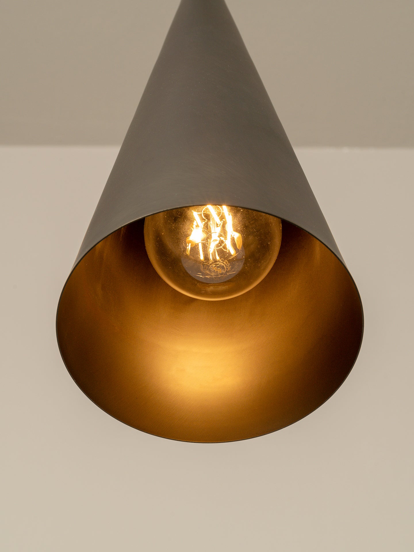 A cone-shaped pendant light fixture with a matte dark gray exterior and a metallic gold interior, showcasing a glowing warm Edison bulb.