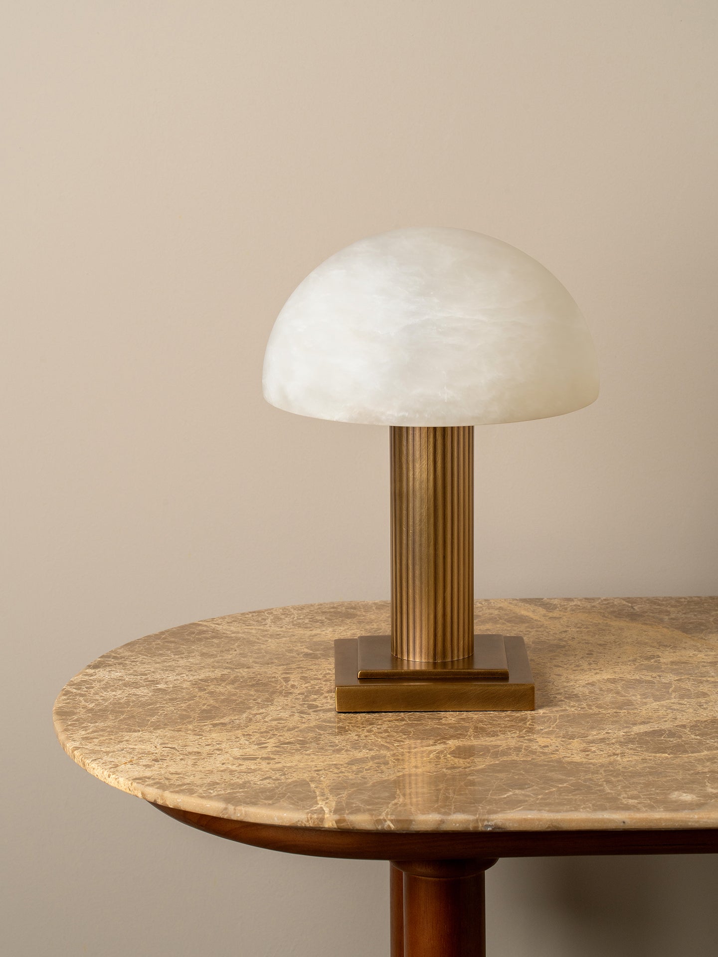 A mushroom-shaped table lamp with a textured white glass dome shade and a fluted brass column on a matching square base, displayed on a round marble-topped table against a beige wall.