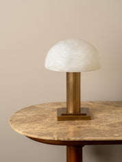 A mushroom-shaped table lamp with a textured white glass dome shade and a fluted brass column on a matching square base, displayed on a round marble-topped table against a beige wall.