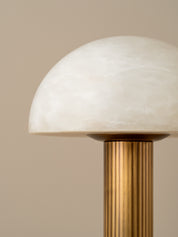 A close-up of a mushroom-shaped table lamp with a white marbled glass dome shade and a fluted brass column on a warm beige background.