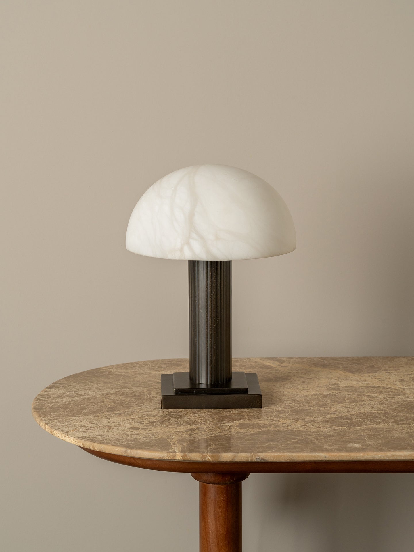 A mushroom-shaped table lamp with a white marble dome shade and a dark bronze cylindrical stem, displayed on a round brown marble table against a neutral beige wall.