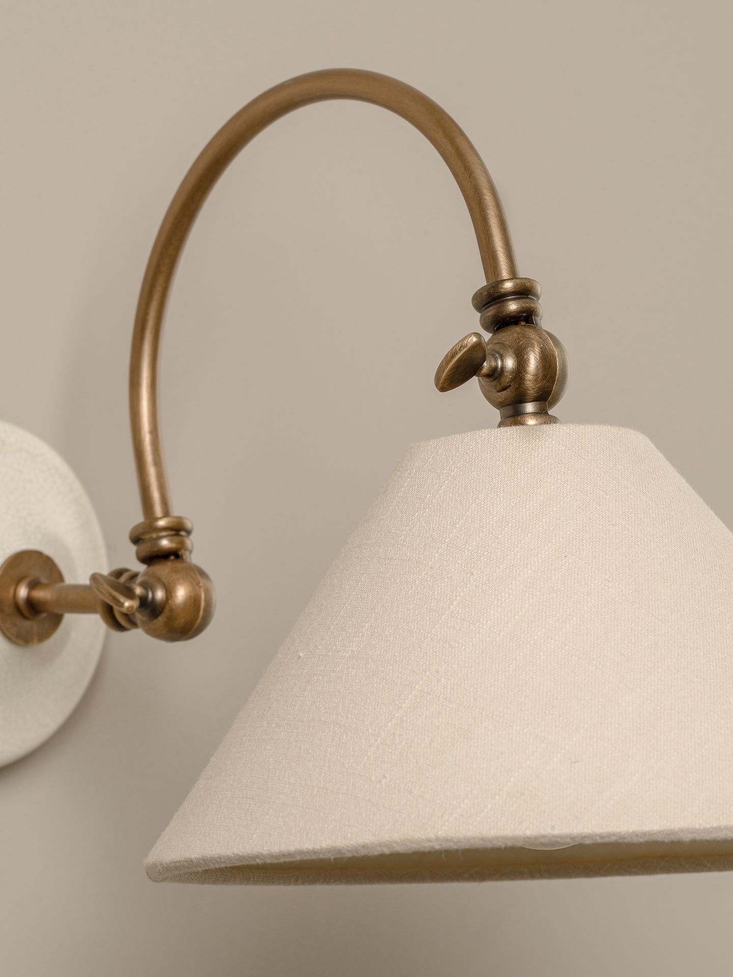 Close-up of a wall-mounted sconce with an arched brass-toned metal arm and a large tapered off-white fabric shade against a warm beige wall.