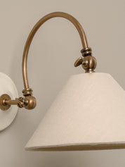 Close-up of a wall-mounted sconce with an arched brass-toned metal arm and a large tapered off-white fabric shade against a warm beige wall.