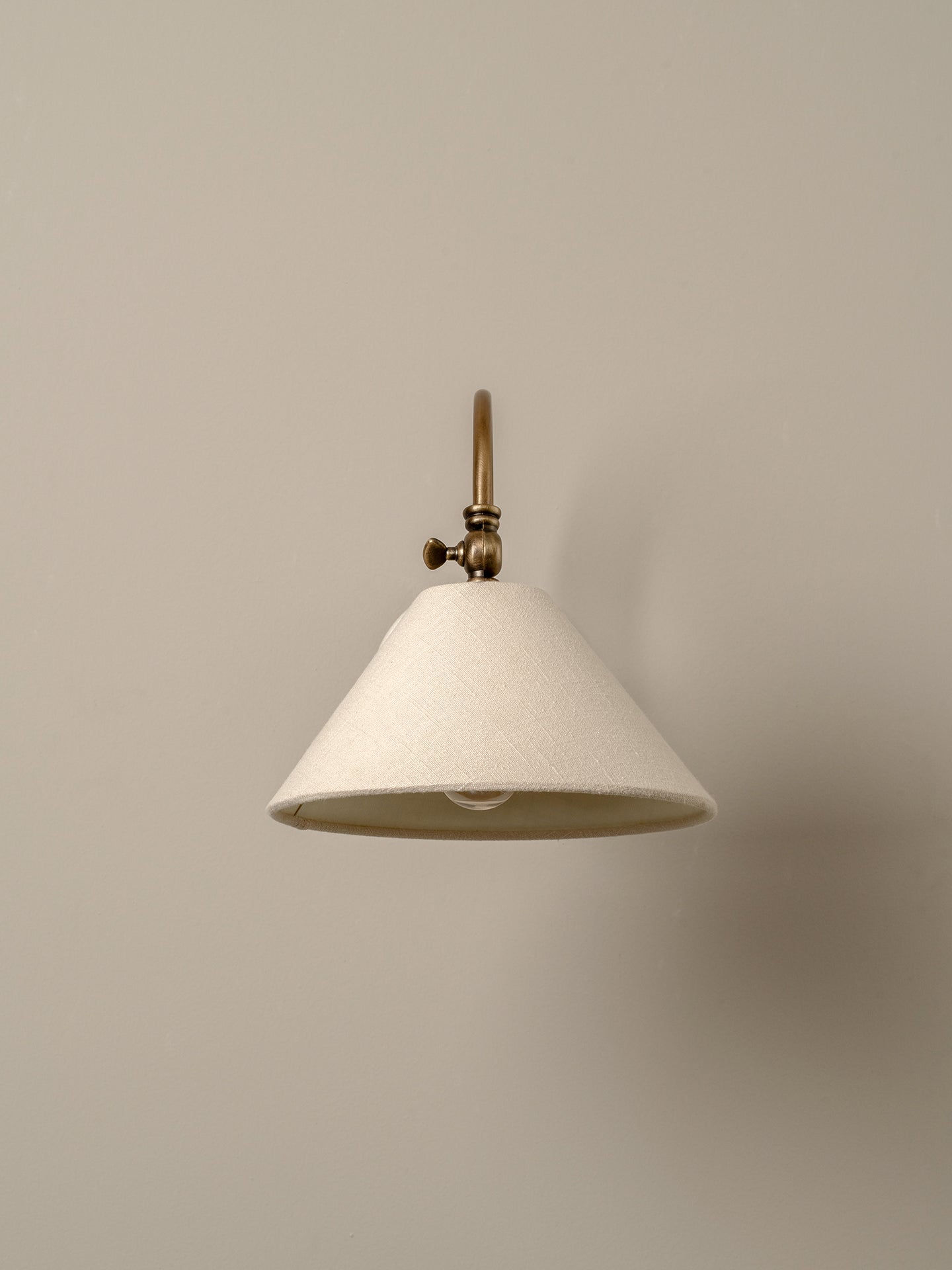 A modern wall sconce with a tapered off-white fabric shade and an antique brass metal fixture, shown against a solid warm beige wall.