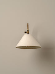 A modern wall sconce with a tapered off-white fabric shade and an antique brass metal fixture, shown against a solid warm beige wall.