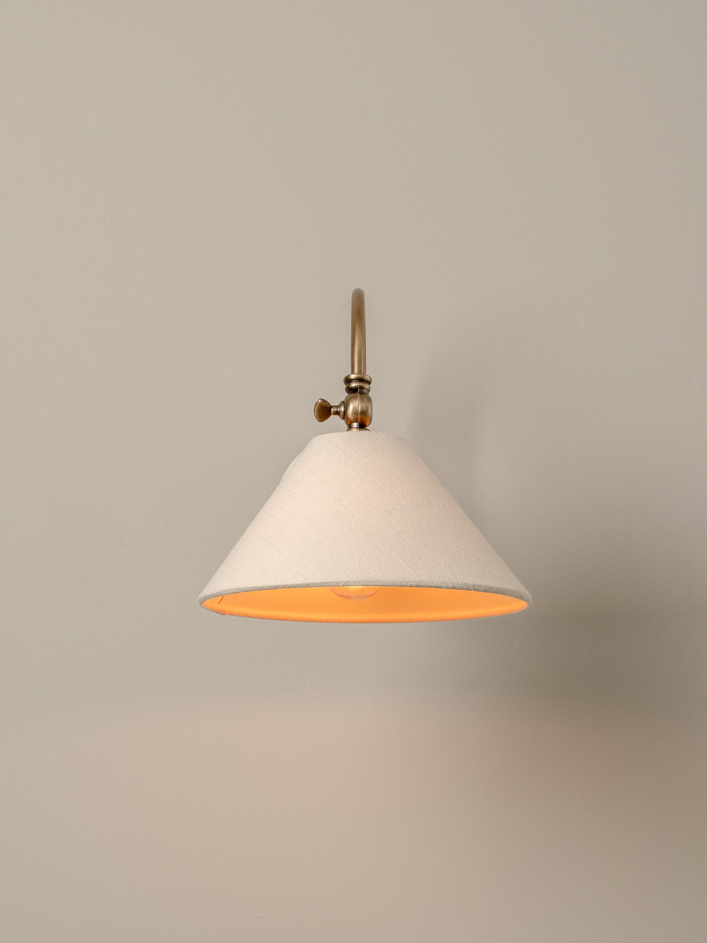 A modern wall sconce with a tapered off-white fabric shade, a warm orange interior, and a brass metal wall mount, shown against a neutral beige wall.