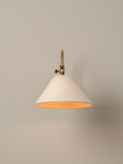 A modern wall sconce with a tapered off-white fabric shade, a warm orange interior, and a brass metal wall mount, shown against a neutral beige wall.