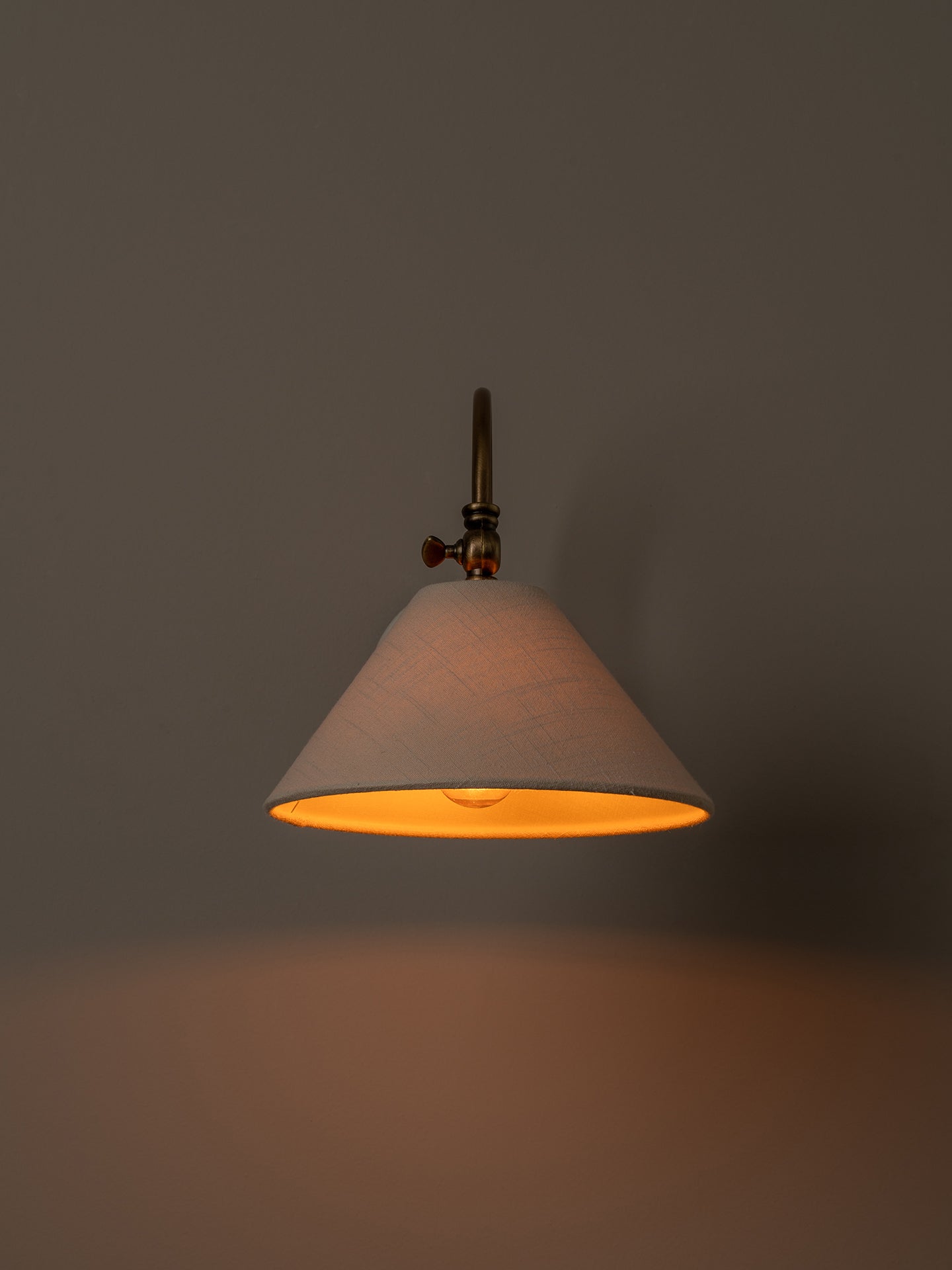 A hanging pendant light with a tapered off-white fabric shade and a warm brass-toned metal fixture, shown illuminated against a dark gray wall.