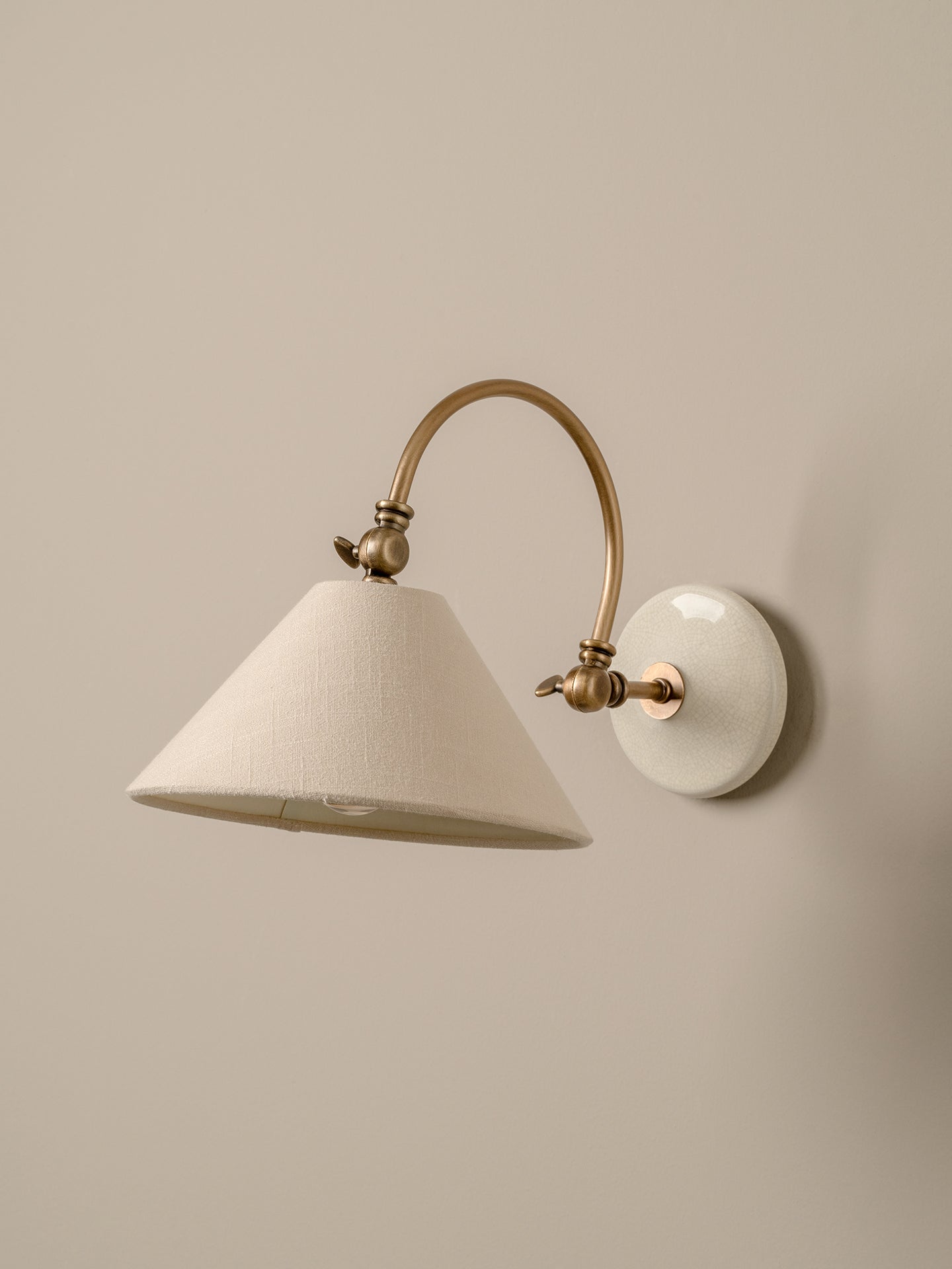 A wall-mounted sconce with a tapered off-white fabric shade, a curved brass arm, and a round white wall plate, shown against a plain beige wall.