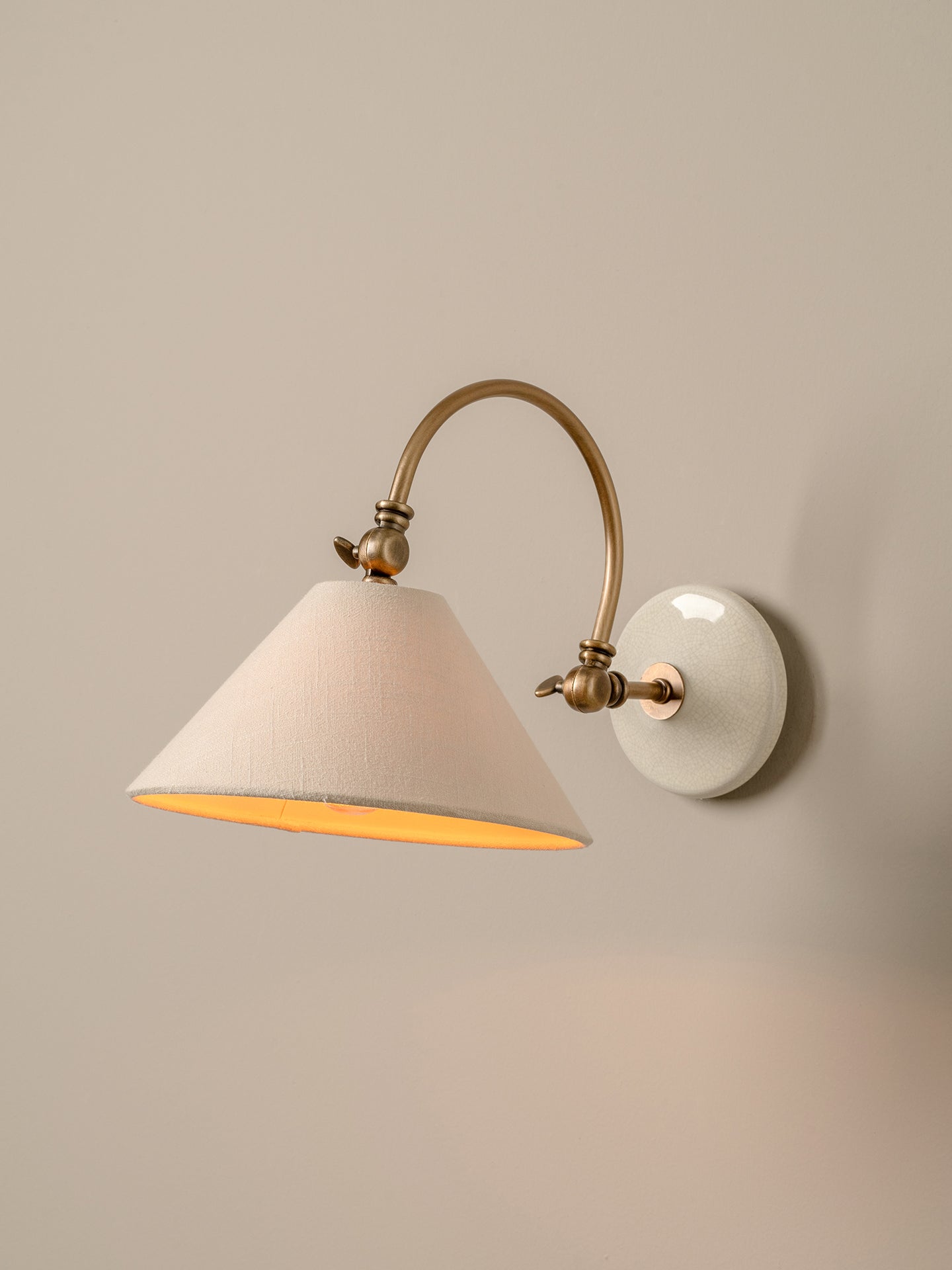 A wall-mounted sconce with a tapered off-white fabric shade, a warm brass curved arm, and a round white ceramic backplate, shown lit against a neutral beige wall.