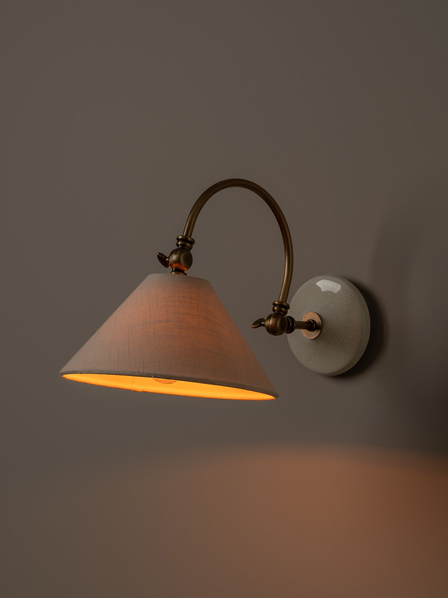 A wall-mounted sconce with a warm glowing conical shade, an arched brass-toned metal arm, and a round white wall plate against a dark gray wall.
