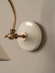 Close-up of a wall-mounted light fixture with an oval white glaze shade and curved brass-toned metal arm against a warm beige wall.
