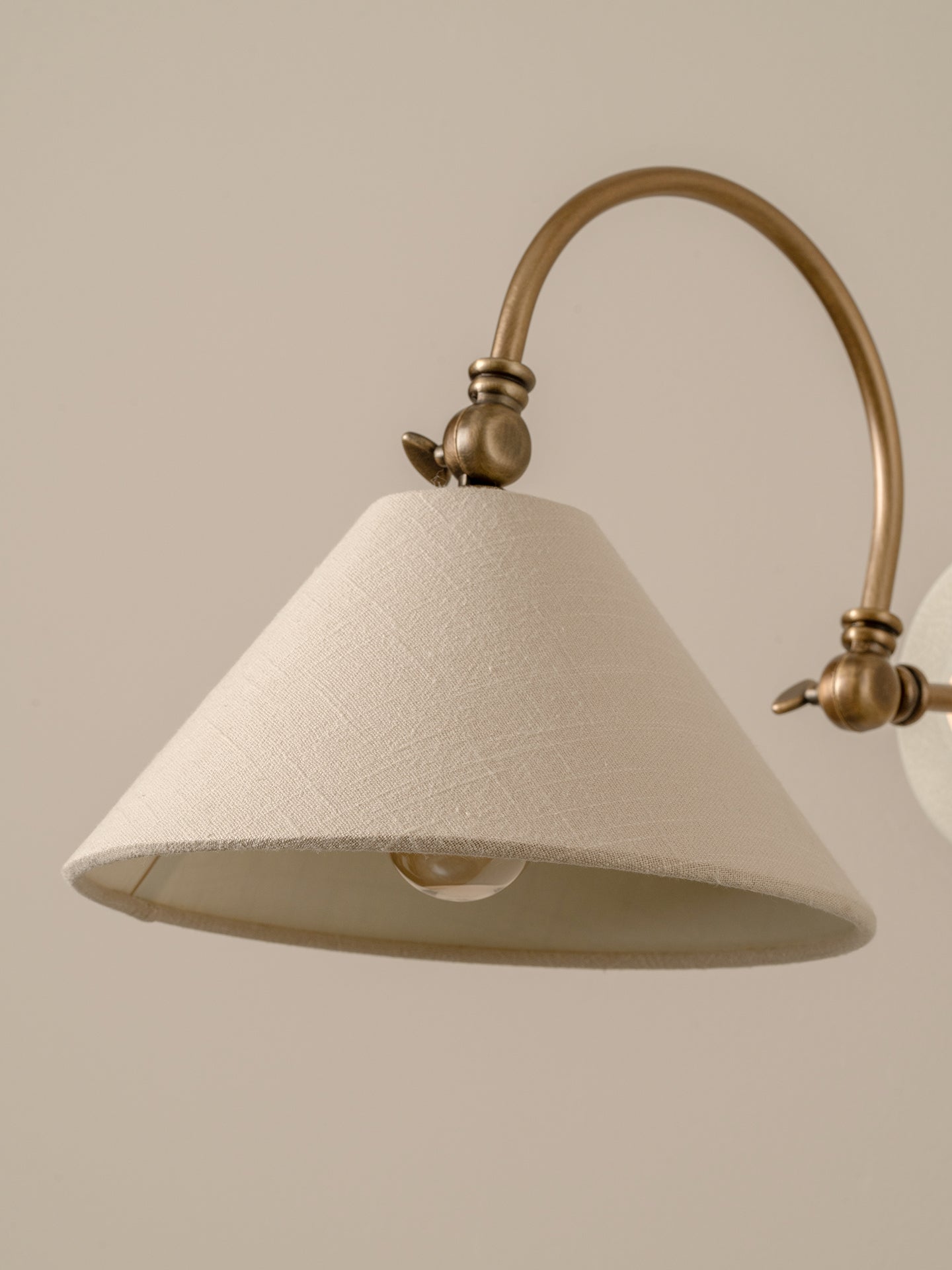 A close-up of a wall sconce featuring a textured off-white fabric shade and an antique brass curved arm against a solid beige background.