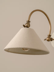 A close-up of a wall sconce featuring a textured off-white fabric shade and an antique brass curved arm against a solid beige background.