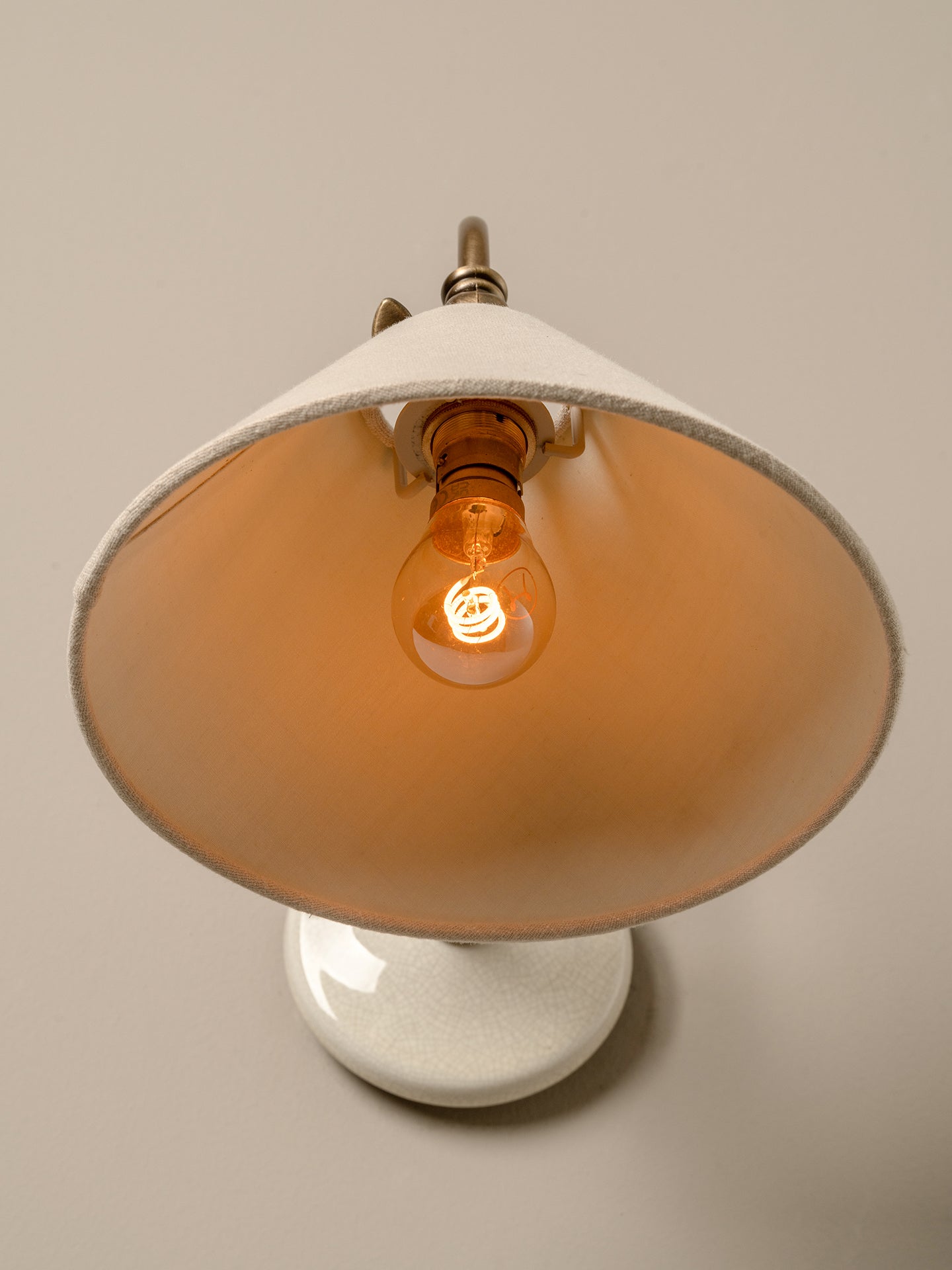 A vintage-style table lamp with a wide, conical off-white shade, a white round base, and a glowing warm amber light bulb, shown against a solid neutral beige background.
