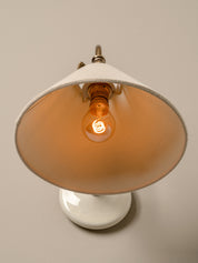 A vintage-style table lamp with a wide, conical off-white shade, a white round base, and a glowing warm amber light bulb, shown against a solid neutral beige background.