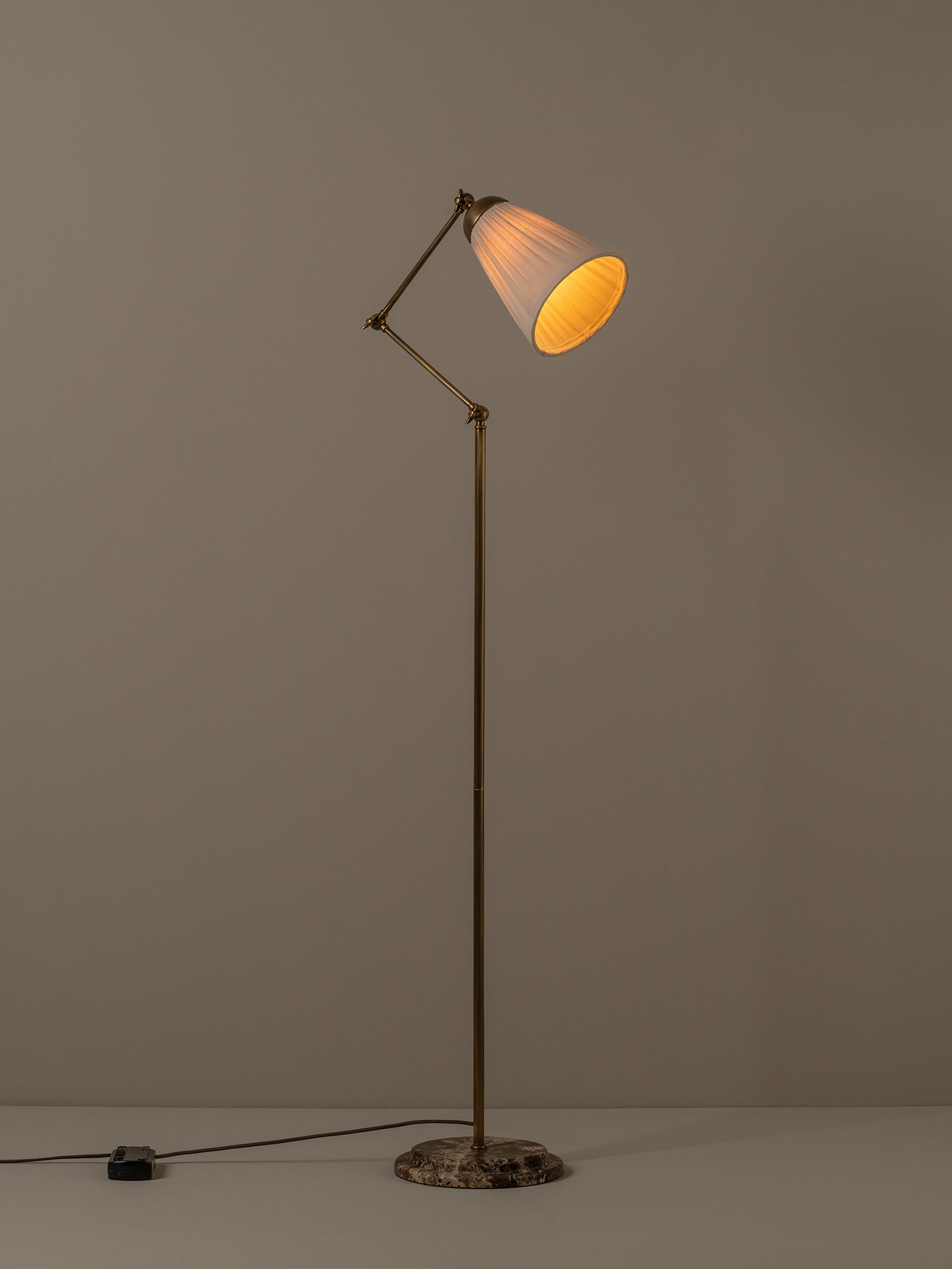 An adjustable floor lamp with a brass-finished metal frame, a pleated white shade, and a round dark marble base, shown lit against a neutral gray background.