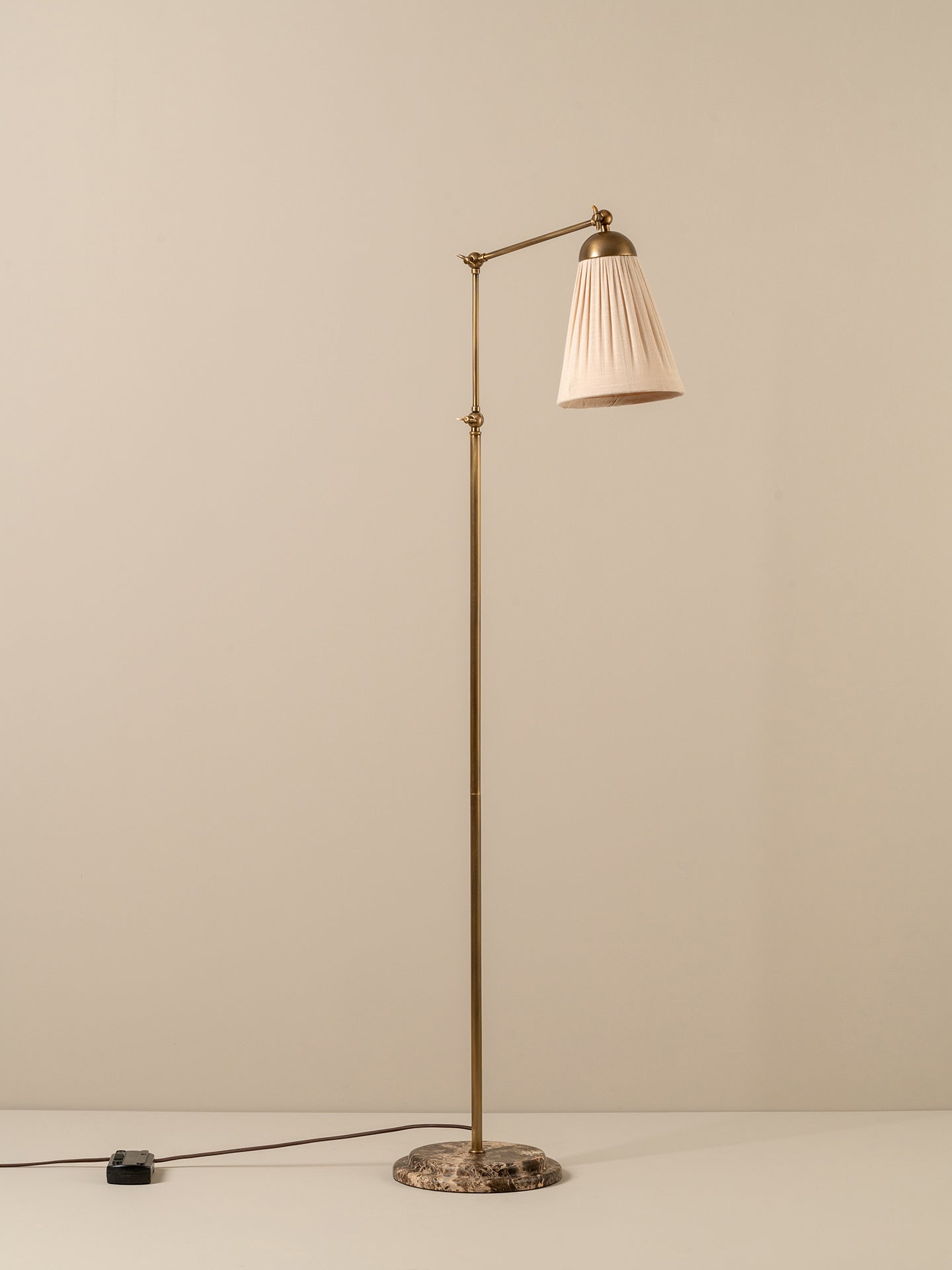 A mid-century modern floor lamp with a brass-colored metal stand, pleated off-white shade, and a round stone base, set against a neutral beige background.