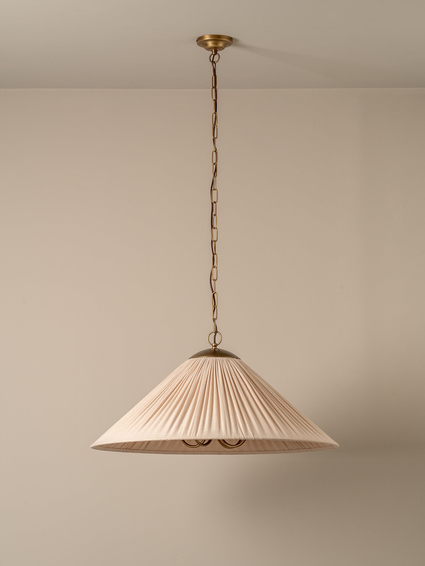 A pleated conical pendant light shade in a warm cream or off-white fabric, illuminated with a soft warm glow. It hangs from a dark brass chain and ceiling canopy against a plain, warm beige wall.