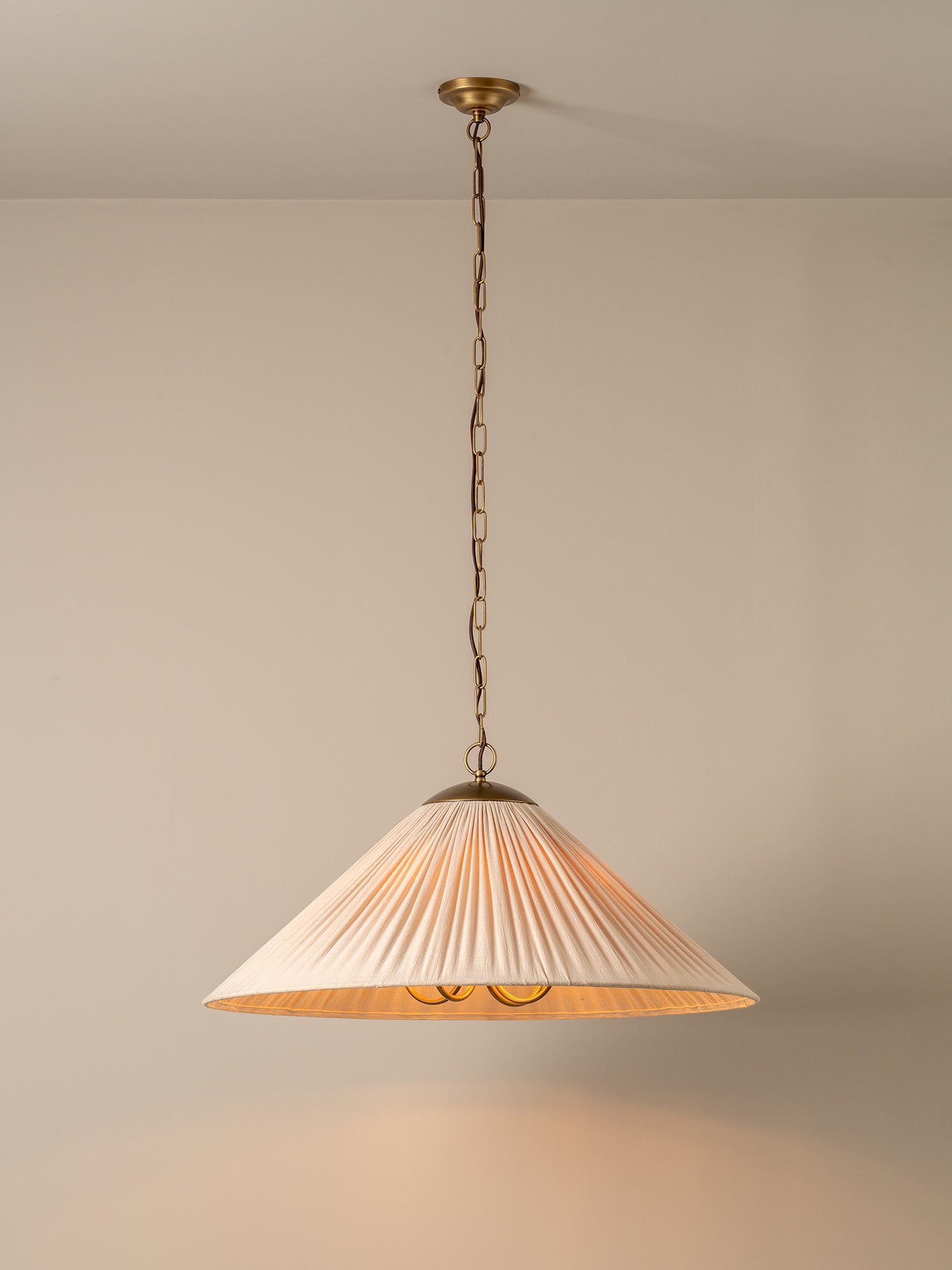 A pleated conical pendant light shade in a warm cream or off-white fabric, illuminated with a soft warm glow. It hangs from a dark brass chain and ceiling canopy against a plain, warm beige wall.