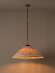 A pleated conical pendant light shade in a warm cream or off-white fabric, illuminated with a soft warm glow. It hangs from a dark brass chain and ceiling canopy against a plain, warm beige wall.