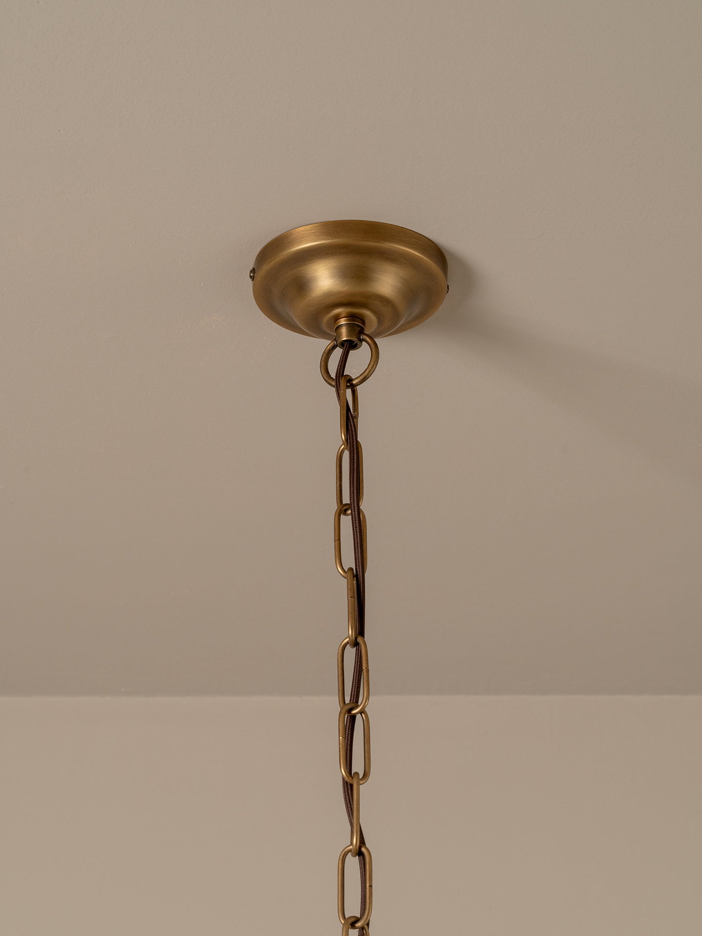 A pleated conical pendant light shade in a warm cream or off-white fabric, illuminated with a soft warm glow. It hangs from a dark brass chain and ceiling canopy against a plain, warm beige wall.